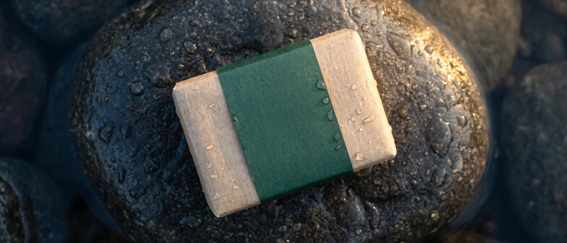A soap bar wrapped in plain kraft paper with a deep green band, resting on wet dark river stone.